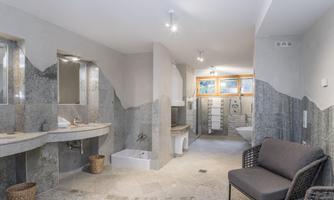 A modern bathroom with gray stone walls and an elegant sink. There is a cozy sitting area and plenty of natural light coming through the windows.