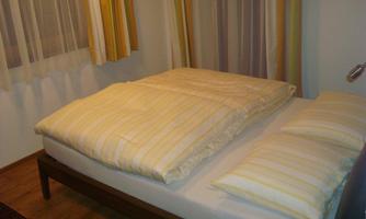 A cozy bedroom with a bed covered with a striped bedspread and pillows. The windows are decorated with light curtains.