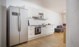 A modern kitchen with a large fridge and a stove. In the background, there is a cozy sofa and a window with curtains.