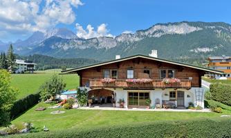 A traditional house with a balcony and flowers, surrounded by a green meadow. In the background, you can see tall mountains and a blue sky.