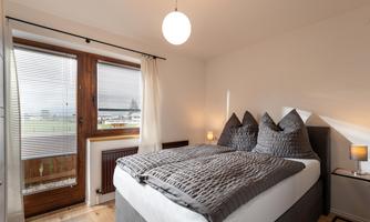 A bright bedroom with a large bed and cozy pillows. Windows overlooking the outdoors provide natural lighting.