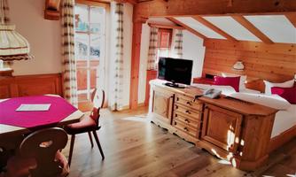 A cozy room with wooden decor, a table and chairs, as well as a television. The window offers a view of the balcony and natural lighting.
