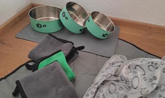 Three feeding bowls and various blankets lie on a floor covering. The bowls are mint green with a paw print pattern.