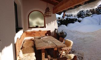 A cozy terrace with a wooden table and chairs, surrounded by freshly fallen snow. A lamp hangs above the table, and snow-covered hills can be seen in the background.