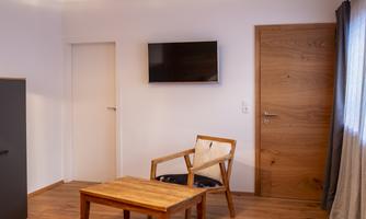 A bright room with a wooden chair and a table. There is a television on the wall as well as a wooden door.