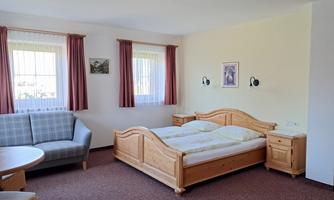 A cozy bedroom with a double bed and a sofa. The room has bright walls and large windows with curtains.