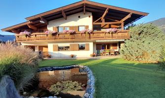 A beautiful house with a large balcony and many flowers. In the foreground, there is a small pond and a lot of greenery.