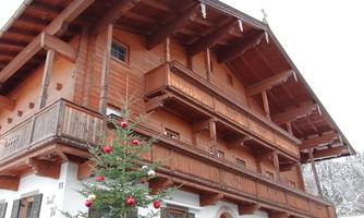 A traditional Alpine wooden house with several balconies. In front of the house stands a Christmas tree adorned with red ornaments.