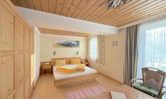 A bright and cozy bedroom with wooden furniture. There is a bed, a table, and a chair, as well as large windows that let in a lot of light.