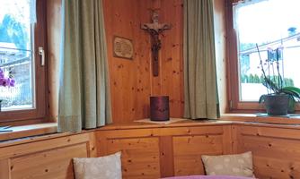 A cozy room with wooden walls and a round table. A cross hangs on the wall, and the windows are adorned with green curtains.