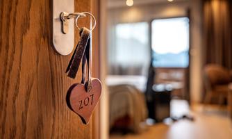 A heart-shaped keychain with the number 207 is hanging on a door. In the background, a cozy room with a bed and chairs can be seen.