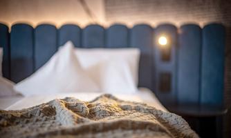 A cozy bedroom with a soft bed and blue headboard. A wool blanket lies on the bedspread and warm light creates an inviting atmosphere.