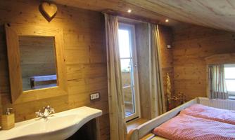 A cozy wooden room with a window and a large mirror. Red cushions are on the bed, and a warm atmosphere is created by the lighting.