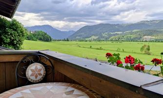 A view from a balcony overlooking green meadows and mountains. Red flowers bloom on the table and surround the scene.