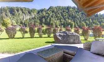 A cozy terrace with modern furniture overlooks a green meadow and wooded hills. The sky is clear and the surroundings exude tranquility.