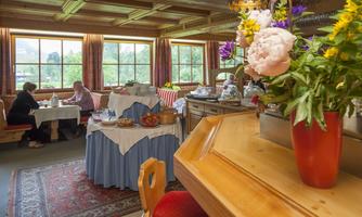 An inviting restaurant with wooden elements and a view of greenery through the windows. Fresh flowers and a variety of dishes are on the table.