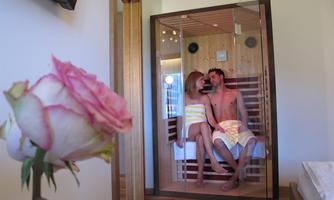 A couple is sitting relaxed in a sauna. In the foreground, there is a pink flower that creates a cozy atmosphere.