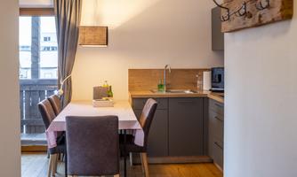 A modern kitchen with a dining area and a wooden chair. A table is set with a white tablecloth and there are windows with curtains.
