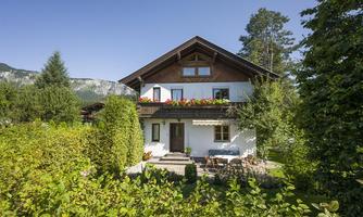 A beautiful house surrounded by a green garden and trees. In the background, gentle mountains can be seen.