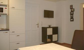 A modern kitchen with white cabinets and a microwave. In the background, there is a TV and a small table.