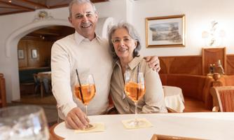 A happy older couple smiles at a bar, holding two cocktails in their hands. The atmosphere is warm and inviting, with cozy decor in the background.