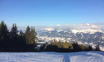 A snowy landscape with mountains in the background and a clear blue sky. In the foreground lies a groomed ski slope.