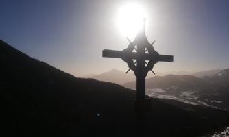 A large cross in front of a majestic mountain landscape. The sun shines brightly in the background, creating a nostalgic atmosphere.