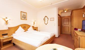 A cozy hotel room with two beds and warm wooden furniture. The room is brightly lit and features a small seating area.