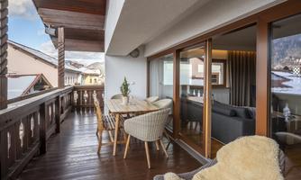 A cozy terrace with a wooden table and comfortable chairs. In the background, alpine houses and a beautiful mountain landscape can be seen.