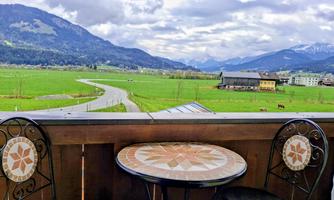A beautiful view from a balcony overlooking green meadows and mountains in the background. Two chairs and a round table are on the balcony.