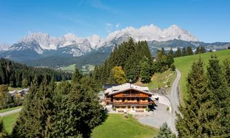 A charming mountain house amidst green meadows and dense forests. In the background, majestic mountains rise up with snow-capped peaks.