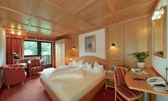 A cozy hotel room with wooden decor and large windows. There is a double bed, a workspace, and comfortable armchairs.
