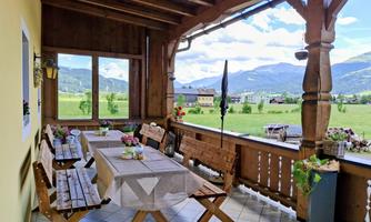 A cozy porch with wooden furniture and a long tablecloth. In the background, green meadows and mountains can be seen.