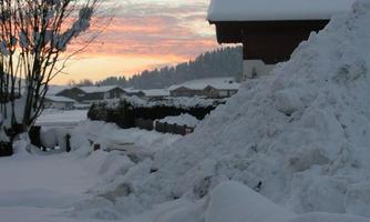 A snowy landscape with a large pile of snow and a house in the background. The sky is painted in soft colors at sunrise.