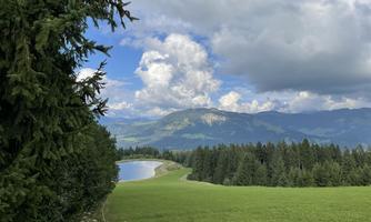 A beautiful landscape with green meadows, a small lake, and forested mountains in the background. The clouds in the sky add a picturesque touch to the scene.