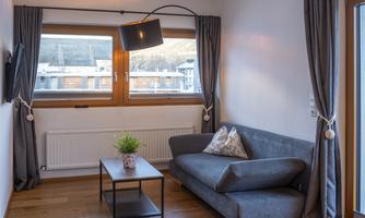 A modern living room with a gray sofa and a small table. Large windows with curtains let in plenty of natural light.