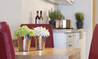 A table with two vases containing green and white flowers. In the background, a modern kitchen with plants and wines can be seen.