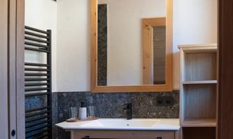 A modern bathroom with wooden shelves and a large mirror. White towels are neatly stored, and the walls are partially covered with dark stones.