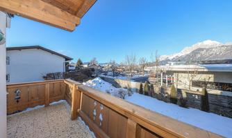 A balcony with a wooden railing, overlooking a snowy landscape. In the background, mountains and snow-covered roofs can be seen.