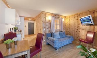 A cozy living space with wooden panels, a blue sofa, and a dining table. A television is mounted on the wall, and there are some plants in the room.