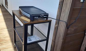 A grill on a cart is standing against the wall. The cart has two levels and wheels for mobility.