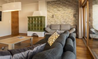 A modern living room with a cozy sofa and a wooden table. A large window view and a green corner with a stove give the room an inviting atmosphere.