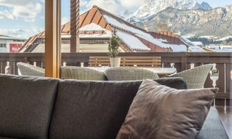 A cozy living room with a gray sofa and a cushion. In the background, you can see snow-covered mountains and a terrace with furniture.