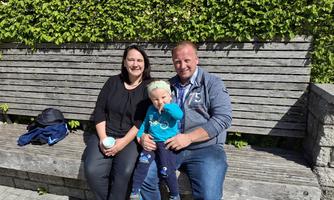 A family sits relaxed on a wooden bench. The father holds a child in his arms while the mother smiles.