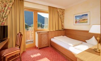 A cozy hotel room with light curtains and a large window. It offers a view of the mountains and is equipped with a single bed and a desk.