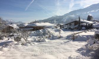 A snowy landscape with houses and snow-covered trees. In the background, the mountains can be seen under a blue sky.