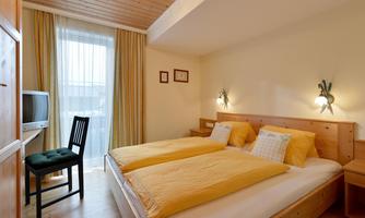 A cozy bedroom with two beds, a television, and a chair. The room has large windows and is brightly designed.