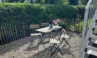 A cozy outdoor area with a table and four chairs. The green hedge and flowers give the scene an inviting atmosphere.