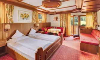 A cozy bedroom with two beds, wooden paneling, and colorful curtains. The room features a small table and a comfortable seating area.