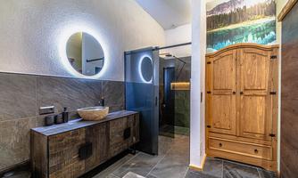 A modern bathroom with a stylish wooden dresser and a round mirror. The walls are clad with tiles, and there is a shower as well as a picturesque wall design.
